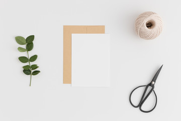 Top view of a white card mockup and envelope with twine, scissors and green branch on a white table.