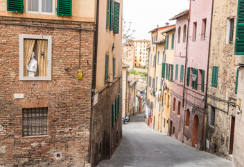 looking down a steep street in siena, italy. a liflike painting n the wall shows a woman looking out the window behind the curtains. Makes people stop and look twice when passing by.