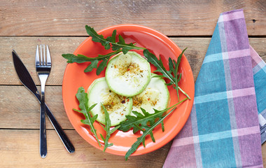 Arugula salad leaves and cooked zucchini with spices in a living coral plate served on wooden rustic table, flat lay, overhead top view, closeup, copy space, vegetarian food concept