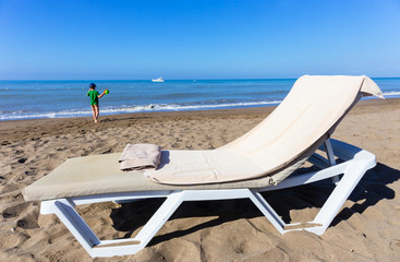 Fototapeta na wymiar Concept of rest by the sea - lounger with beach towel and child walking to sea