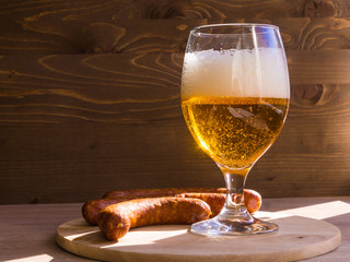 glass of beer with foam head, illuminated by bright sunshine and a smoked sausage on a dark wooden background