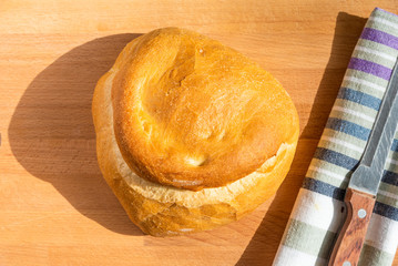 A loaf of homemade wheat bread on a wooden board, a rustic striped kitchen towel and a knife with a wooden handle, top view, natural light