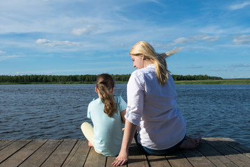 Fototapeta premium mom and daughter sit on the pier and look at the beautiful lake and nature