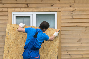 a man obstructs a window with a large piece of plywood before a natural disaster, a hurricane