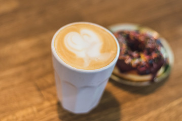 coffee with a heart made of milk and a donut on a wooden table. Coffee with a drawn heart and milk on a wooden table in a coffee shop. chocolate donut with scattering on the table next to the coffee
