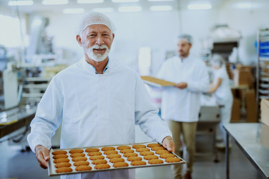 Food plant workers in sterile uniforms carrying trays with cookies. Selective focus on senior adult worker.