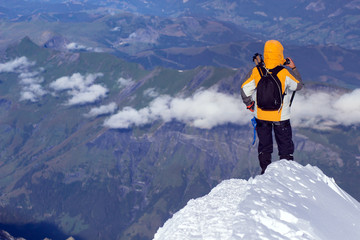 Man in a yellow jacket with a hood standing on a rock with his hands up and looking at the sunset in the mountains, a back view
