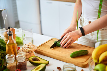 Obraz na płótnie Canvas Young happy woman cutting cucumber for making salad in the beautiful kitchen with green fresh ingredients indoors. Healthy food and Dieting concept. Loosing Weight