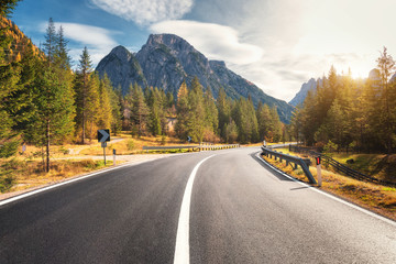 Road in summer forest at sunset in Italy. Beautiful mountain roadway, trees with green foliage and sunlight. Landscape with empty asphalt road through woodland, blue sky, high rocks. Travel in Europe