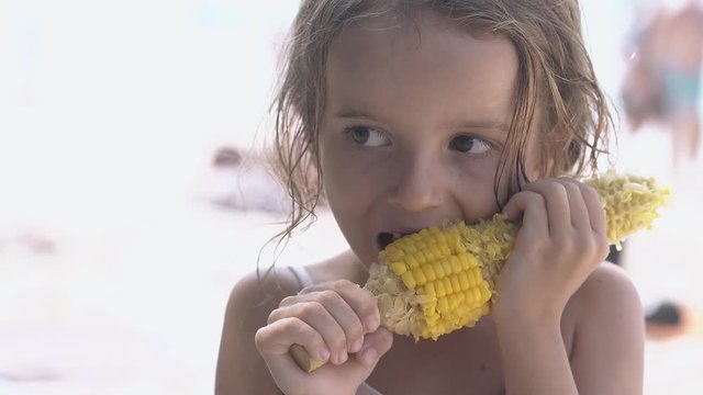 cute little girl with big brown eyes stands on beach and gnaws big corncob on hot day close view