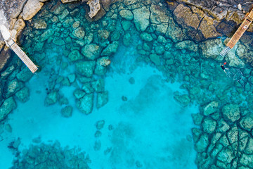 An aerial view of the beautiful Mediterranean Sea, with two wooden piers and a rocky shore, where you can see the textured underwater corals and the clean turquoise water of Protaras, Cyprus