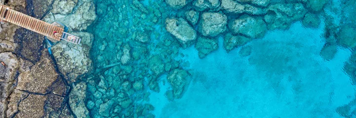 Fotobehang Een luchtfoto van de prachtige Middellandse Zee, met een houten pier en een rotsachtige kust, waar je de getextureerde onderwaterkoralen en het schone turquoise water van Protaras, Cyprus kunt zien © Valentinos Loucaides