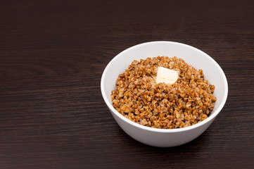 Buckwheat porridge with butter in a small white plate. Buckwheat porridge on a brown background. Bowl with boiled buckwheat. Dietary breakfast. Healthy food
