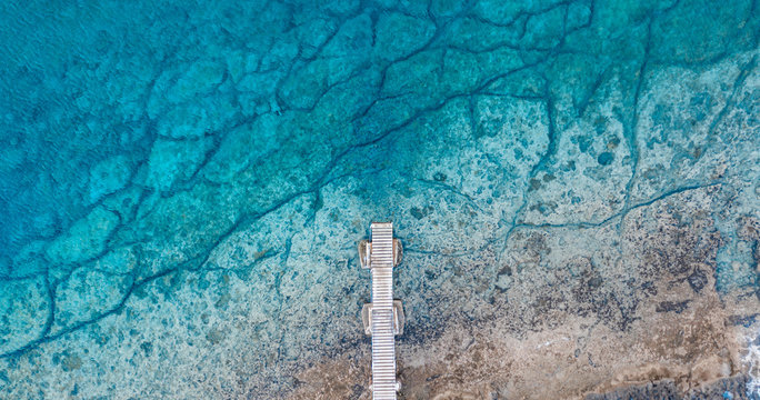 An aerial view of the beautiful Mediterranean Sea, with a wooden pier and a rocky shore, where you can see the textured underwater corals and the clean turquoise water of Protaras, Cyprus