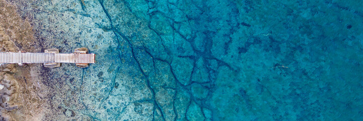 An aerial view of the beautiful Mediterranean Sea, with a wooden pier and a rocky shore, where you can see the textured underwater corals and the clean turquoise water of Protaras, Cyprus