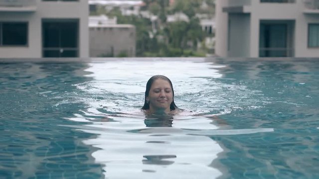 slim tanned lady dives swooping down into crystal blue water against big tropical resort hotel and lush green trees