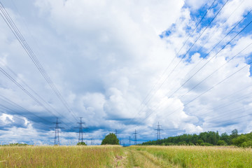 Towers of electric main with the wires in the summer countryside field on the background of blue sky with clouds and the forest 