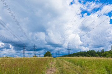 Fototapeta na wymiar Towers of electric main with the wires in the summer countryside field on the background of blue sky with clouds and the forest 