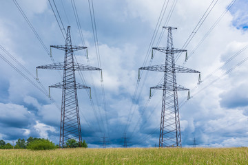 Towers of electric main with the wires in the summer countryside field on the background of blue sky with clouds and the forest 