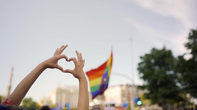 Supporting hands make heart sign and wave in front of a rainbow flag flying on the sidelines of a summer gay pride parade