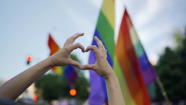 Supporting hands make heart sign and wave in front of a rainbow flag flying on the sidelines of a summer gay pride parade