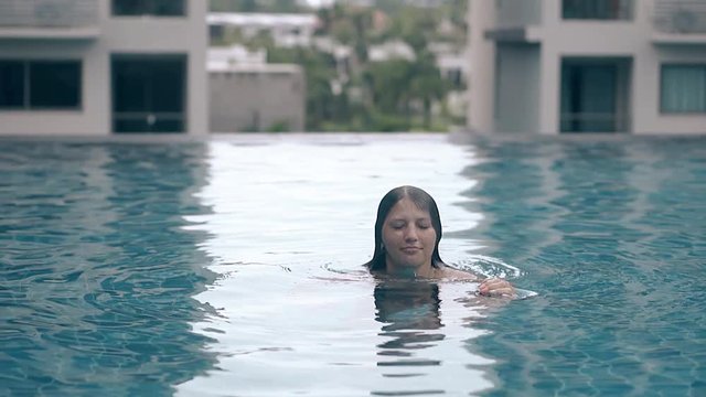 graceful young woman emerges from azure water in tropical hotel roof pool on sunny day slow motion