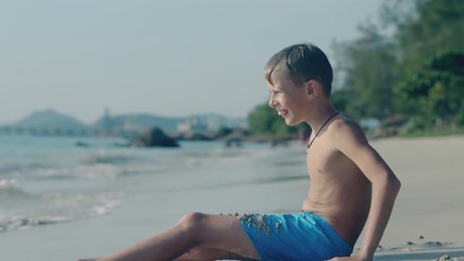 joyful boy in blue shorts sits by ocean edge and throws wet sand balls into water on pictorial beach against clear sky