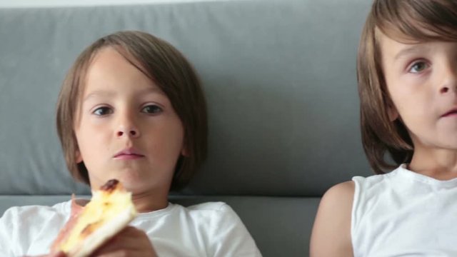 Cute children, sitting on couch, eating pizza and watching TV. Hungry child taking a bite from pizza on a pizza party day at home