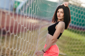 Beautiful stylish brunette woman in black top and pink shorts stands near a football goal at the stadium at sunset. Beautiful sunlight. A girl with a perfect figure and in great shape