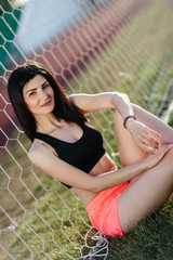 portrait of Beautiful brunette woman in black top and pink shorts sitting on the lawn near a football goal at the stadium at sunset. Beautiful sunlight. A girl with a perfect figure and in great shape