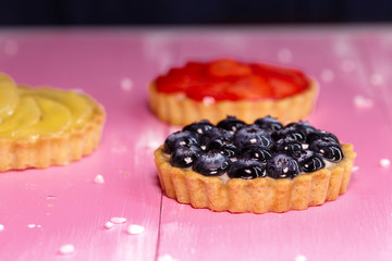 Freshly baked tartlets with strawberry, banana, blueberry fruit filling on a pink wooden background