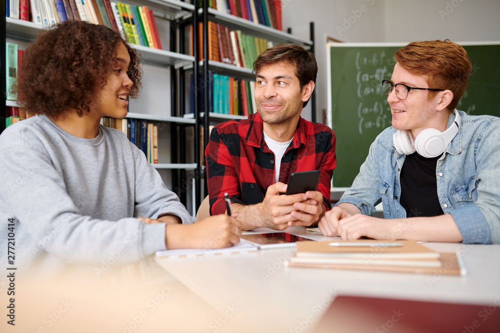 Poster group of teens sitting by table in college library while discussing new ideas