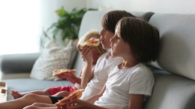 Cute children, sitting on couch, eating pizza and watching TV. Hungry child taking a bite from pizza on a pizza party day at home