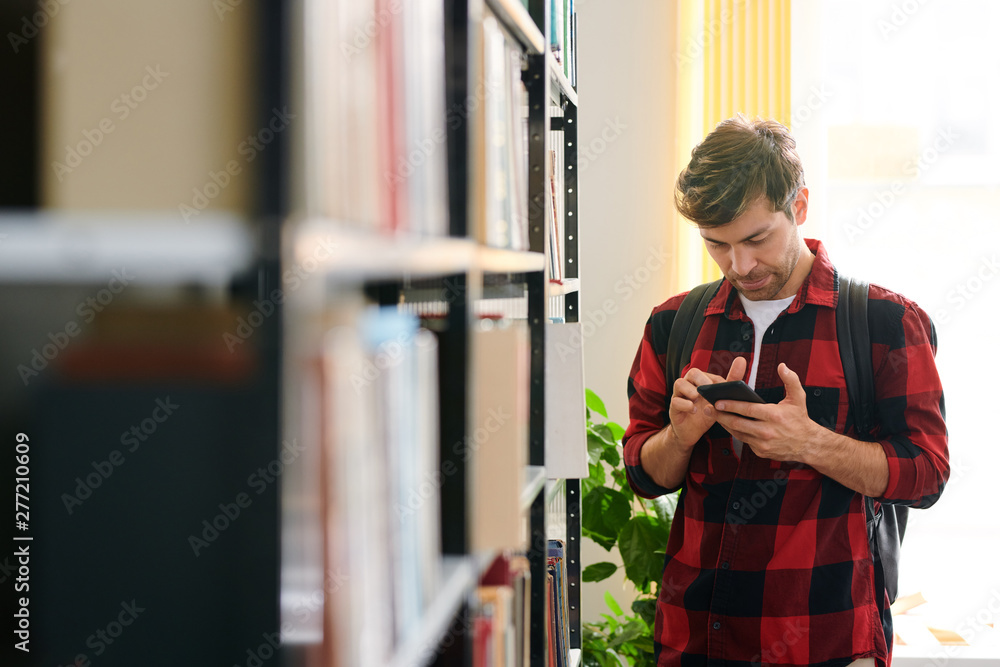 Poster young man scrolling in smartphone by shelf in library
