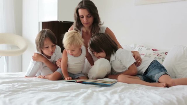 Young mother, read a book to her three children, boys in sunny bedroom at home