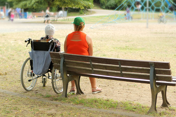 woman in an orange jacket and an old woman are disabled in a wheelchair, sit together in a park in summer
