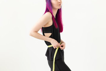 Unidentified young girl hipster with purple hair measures her waist with a measuring tape on a white background. Healthy lifestyle concept. Advertising space.