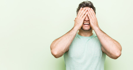 Young handsome man against a green background covers eyes with hands, smiles broadly waiting for a surprise.