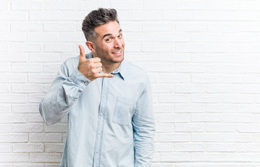 Young handsome man against a bricks wall showing a mobile phone call gesture with fingers.