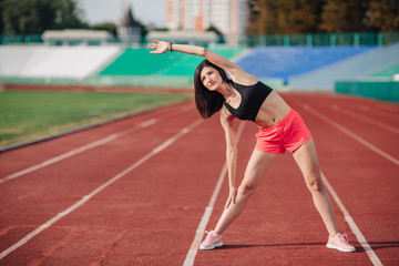 Athlete Attractive sporty brunette woman in pink shorts and top exercise stretching at sport stadium in evening, sunset. Sport and healthy lifestyle