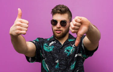 Young man wearing a vacation look showing thumbs up and thumbs down, difficult choose concept