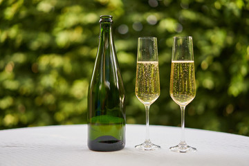 Two wine glasses flute type with sparkling wine or champagne and traditional design champagne bottle on the table with white tablecloth in the romantic garden restaurant ready to celebrate or cheers.
