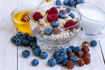 homemade muesli or granola with milk, honey, fresh berries and nuts. healthy muesli with raspberry, blueberry, hazelnut, milk and honey on white wooden rustic table.