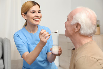 Nurse feeding elderly man with yogurt indoors. Assisting senior people