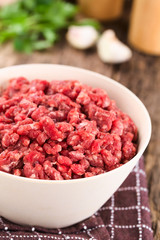 Fresh raw ground or minced beef meat in bowl, seasonings (salt, pepper), garlic and fresh parsley in the back (Selective Focus, Focus one third into the meat)