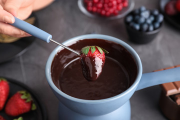 Woman dipping strawberry into pot with chocolate fondue at table, closeup