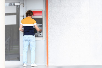 Young man using cash machine outdoors. Space for text