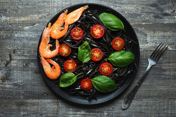 Black pasta (with black cuttlefish ink), cherry tomatoes, shrimps and basil on a black plate. Gray wooden background