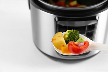 Spoon with vegetables and blurred view of modern multi cooker on background, closeup. Space for text