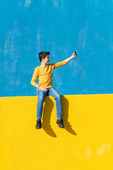 Front view of a young boy wearing casual clothes sitting on a yellow fence against a blue wall while taking a selfie with a smartphone